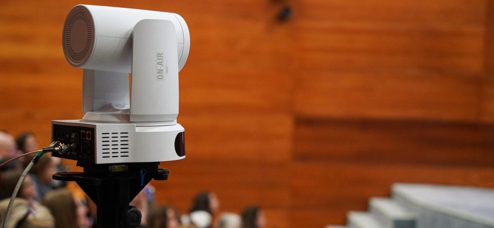 Cámara de vídeo robotizada blanca sobre trípode en un auditorio con público